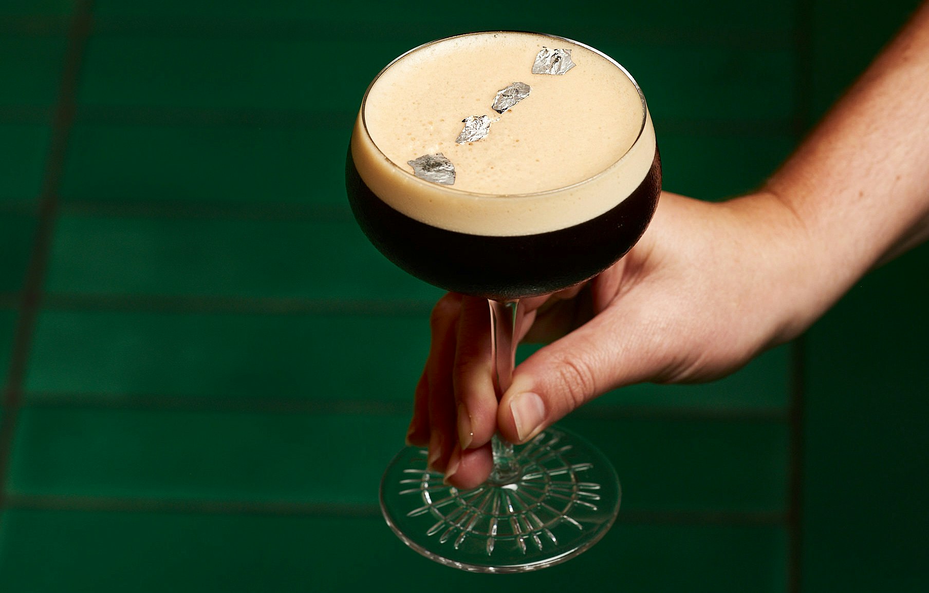 An espresso martini garnished with silver leaf stands on a green table.